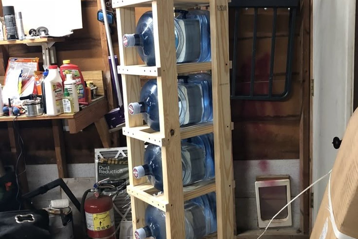 Garage shelf with stacked gallon water jugs, a battery lantern, and labeled fuel cans in soft autumn light.
