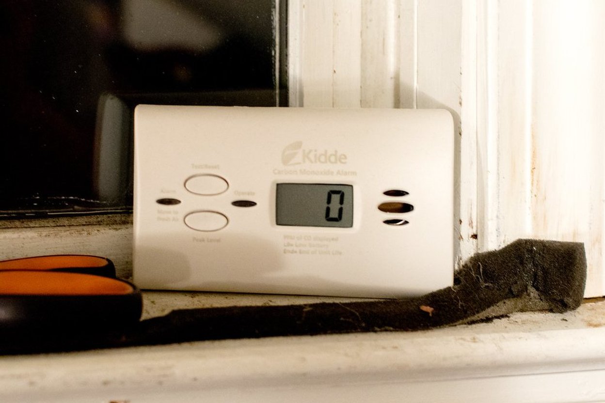 A battery-operated carbon monoxide alarm mounted on a hallway wall outside a bedroom door, with a person pressing its test button.