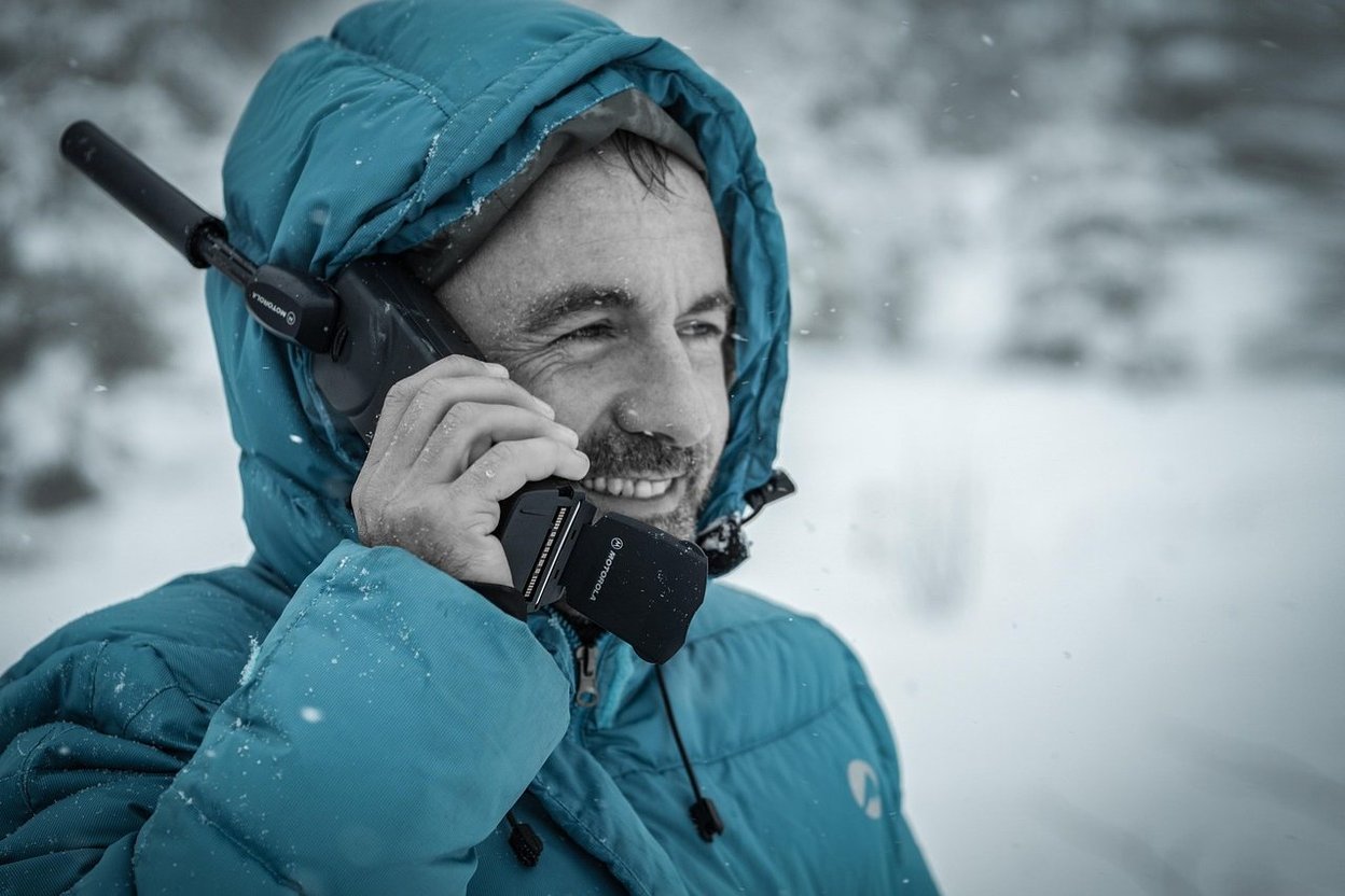 A man in a winter coat talking on a radio in the snow