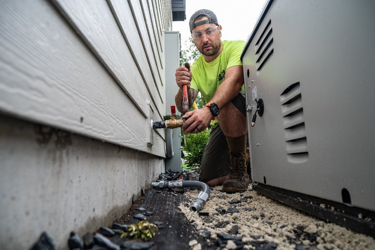 A gray air-cooled home standby generator on a concrete pad beside a brick house, with fallen leaves on the lawn and the gas line and electrical conduit visible